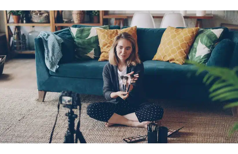 A woman sits on the floor in front of a camera, holding makeup brushes, in a cozy living room with a teal sofa and colorful cushions, influencer, vlogger, beauty