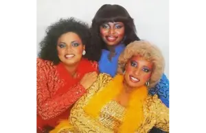 Three women in colorful, glittery outfits. One in red, another in blue, and the third in gold with a feather boa. They are smiling warmly. The one in Yellow is Gwendolyn “Blondy” Chisolm.
