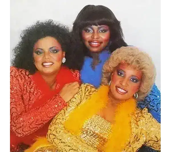 Three women in colorful, glittery outfits. One in red, another in blue, and the third in gold with a feather boa. They are smiling warmly. The one in Yellow is Gwendolyn “Blondy” Chisolm.