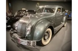 A vintage Chrysler Airflow, a car with a sleek, curved body in a glossy gray finish is displayed in a museum setting. Its design features chrome accents and whitewall tires.