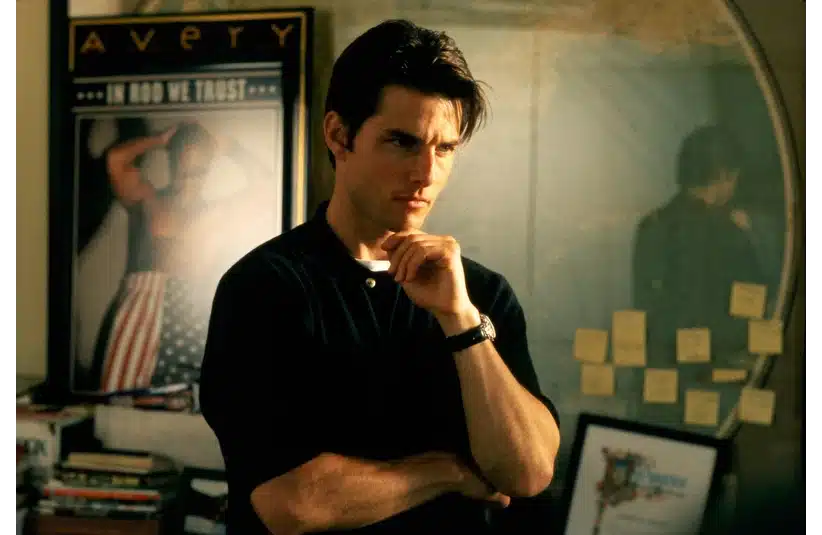 Jerry Maguire, in a thoughtful pose, stands in an office, surrounded by books and sticky notes. A poster and a round mirror are visible in the background.