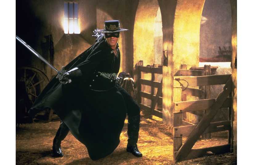 The masked hero, Zorro, played by Antonio Banderas in a black costume and hat, brandishes a sword in a dimly lit stable. The scene is dramatic, evoking a sense of adventure and mystery.