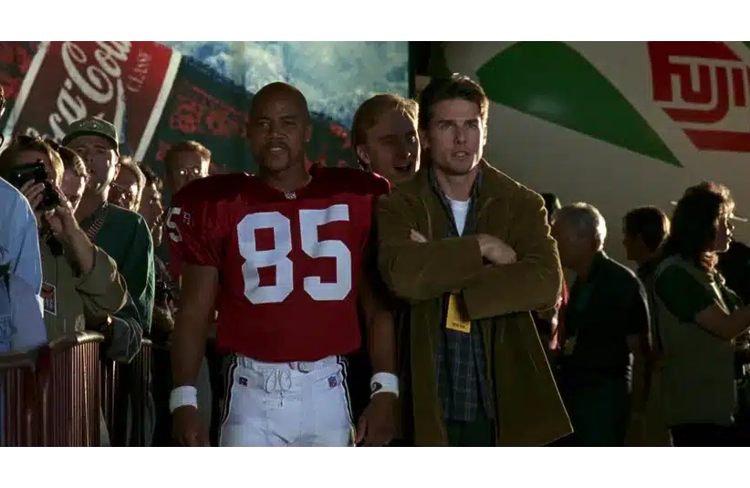 A football player in a red jersey numbered 85 stands confidently next to a man with crossed arms. They're surrounded by a cheering crowd.