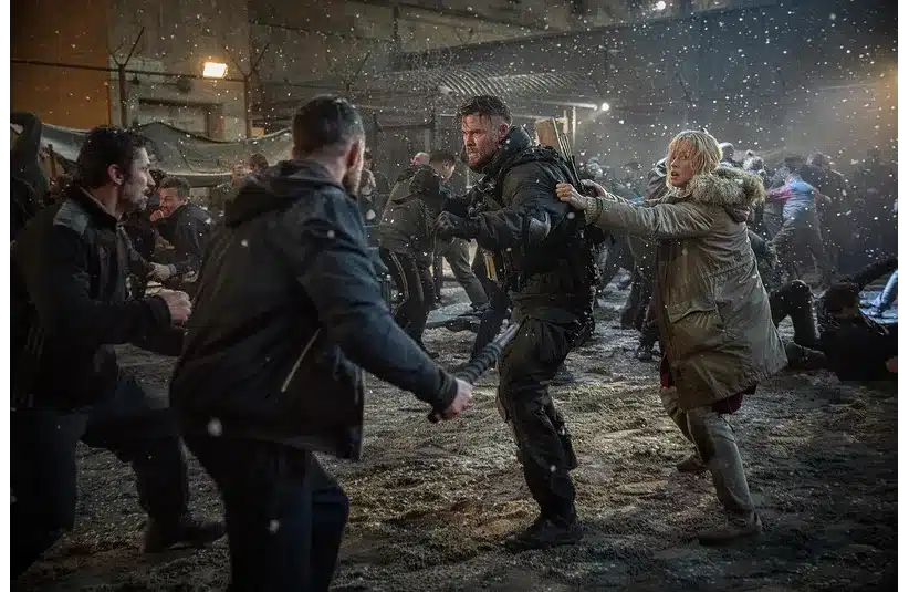 Chaotic scene in a snowy courtyard, with people fighting. A focused man in tactical gear defends a woman, conveying urgency and tension.