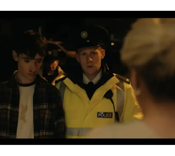 Michael Patrick MND -A young man in a plaid shirt stands at a doorway next to a police officer in a yellow jacket. The scene is tense and dimly lit, conveying urgency.