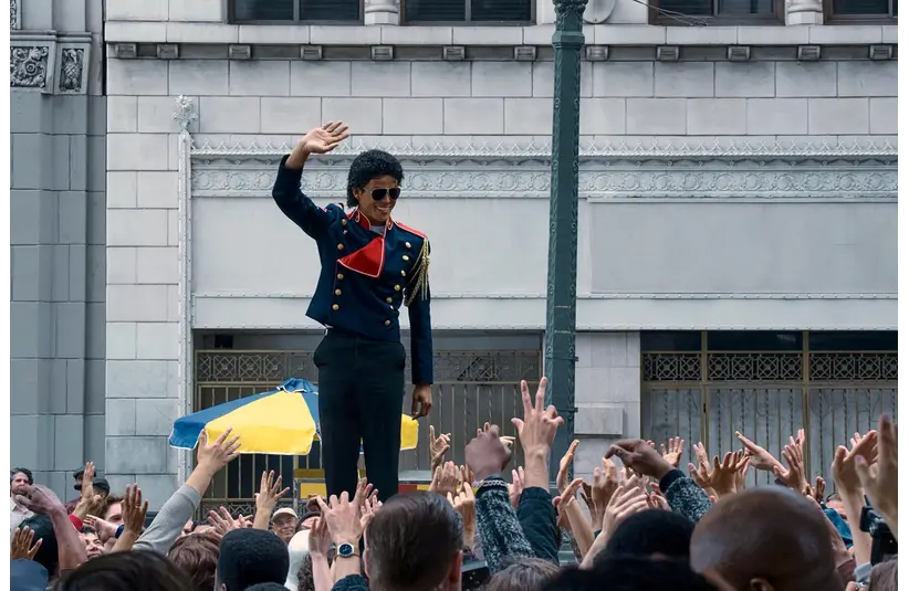 Jaafar Jackson in 'Michael' (2026), in a military-style jacket, waves from a platform above an excited crowd reaching upl. The scene is set against an urban backdrop with a lively, celebratory tone.