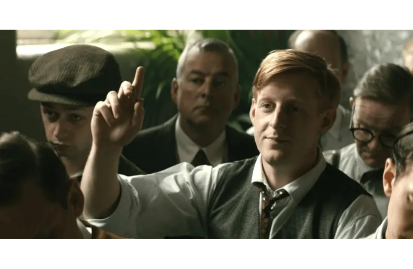 A man with red hair, dressed in a vintage outfit, raises his hand in a meeting. The room is filled with attentive people, creating a serious atmosphere.