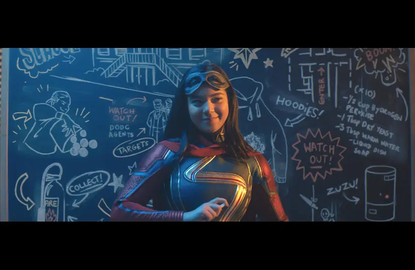 Kamala Khan in Ms. Marvel costume stands confidently in front of a chalkboard filled with doodles and plans. The scene conveys a sense of adventure.