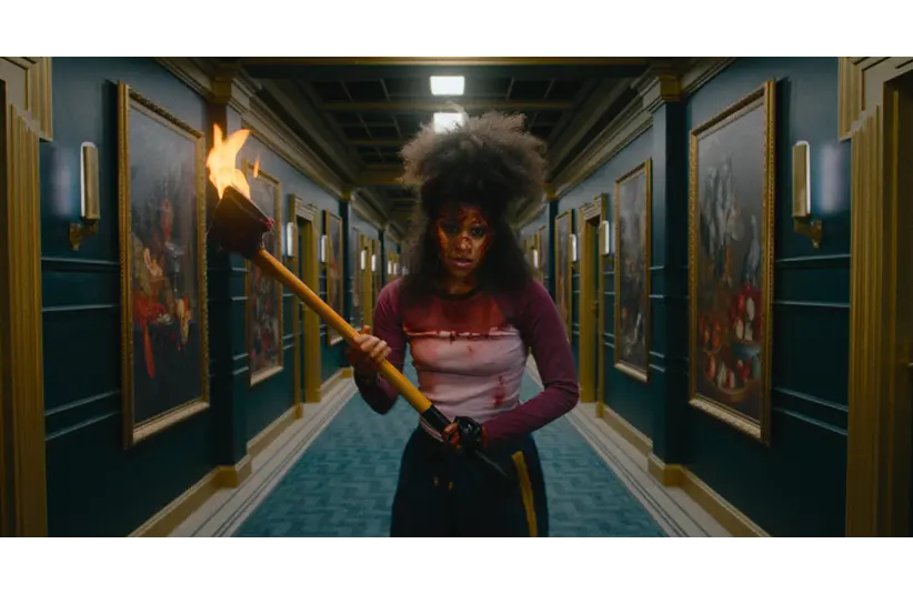 Zazie Beetz with wild hair and face paint stands in a dim hallway holding a burning torch. Framed paintings line the walls, creating a tense, dramatic ambiance.
