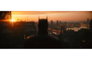 Silhouetted figure with pointed ears stands on a rooftop, gazing over a cityscape at sunset. Buildings and a river glow with warm light. Atmospheric mood. "The Batman: Part 2" is set for release in 2027.