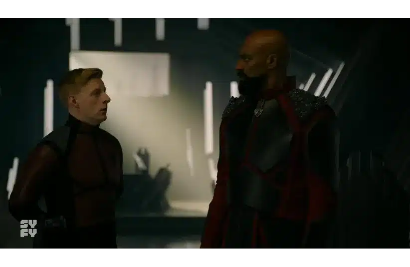 Two characters stand in a dimly lit futuristic room, wearing dark, sci-fi themed costumes. They appear to be in a serious conversation. The atmosphere is tense.