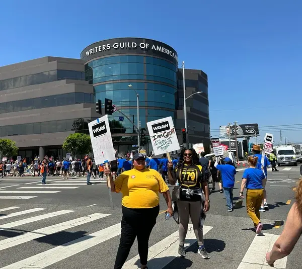 Protesters gather outside the WGA building, holding signs under a clear sky. The atmosphere is determined and unified.