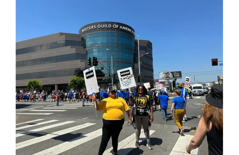 Protesters gather outside the WGA building, holding signs under a clear sky. The atmosphere is determined and unified.