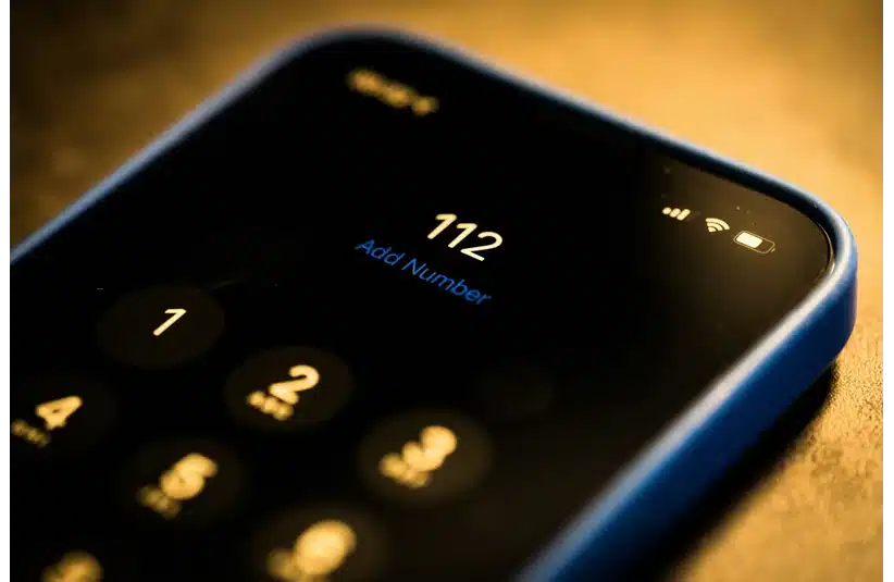 A smartphone screen displaying the emergency number "112" is illuminated in dim lighting. The setting conveys urgency and focus, with a blue phone case visible.