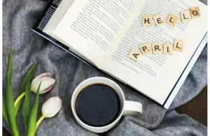An open book with the words "Hello April" spread out across the pages, with a cup of coffee and some tulips on a table.