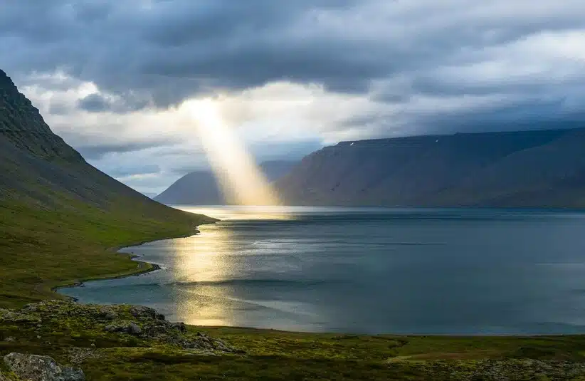 A beam of sunlight pierces through dark clouds, illuminating a serene fjord in Iceland and its surrounding green mountains, creating a peaceful and mystical atmosphere. The Nature of God. Heaven.