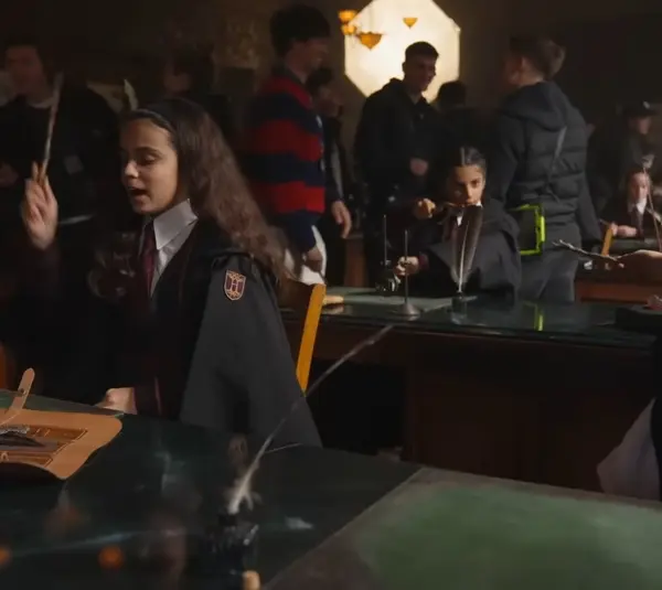 Students in robes practice magic at desks with wands in a dimly lit classroom. The atmosphere is lively and focused, suggesting a magical lesson.