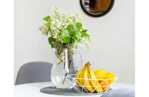 A vase with white flowers and green leaves sits on a round table next to a wire fruit basket filled with bananas and oranges, creating a fresh, inviting atmosphere. Spring decorating