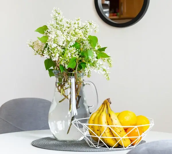A vase with white flowers and green leaves sits on a round table next to a wire fruit basket filled with bananas and oranges, creating a fresh, inviting atmosphere. Spring decorating