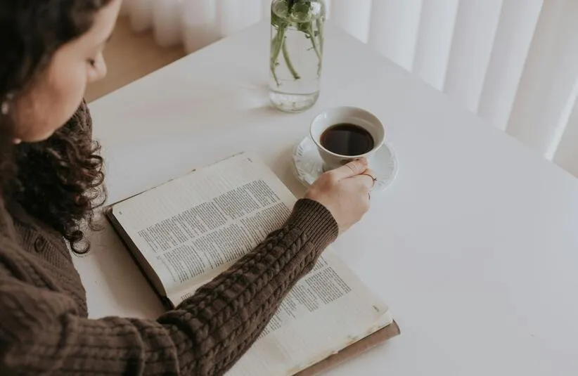 In the daily devotional, a young woman sits with an open Bible and coffee reading.
