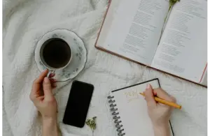 A person writes "Pray" in a notebook on a quilted surface for a daily devotional, next to an open book, a cup of coffee on a saucer, and a smartphone. The scene is calm and reflective.