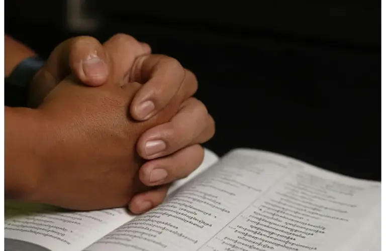 In daily devotion, a person's hands folded in prayer over a Bible are shown.