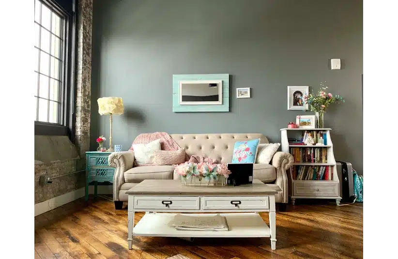 A cozy living room with a beige sofa adorned with pink and blue pillows. A teal side table and floor lamp sit to the left, with a bookshelf and vibrant flowers on the right. A coffee table with pink flowers centers the space, with artwork decorating the gray walls, creating a welcoming atmosphere. Spring decor