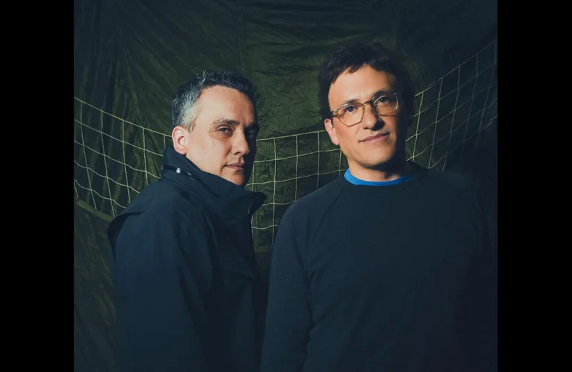 Anthony Russo and Joe Russo, directors of Avengers: Doomsday, stand in front of a green, net-like backdrop. One wears a black jacket, the other a dark sweater, both with neutral expressions.