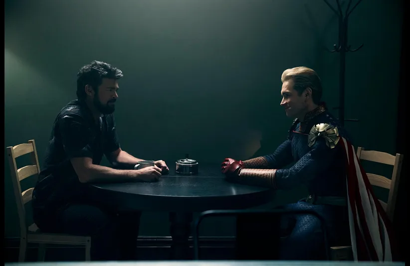 Karl Urban (Billy Butcher) and Antony Starr (Homelander) in The Boys sit across from each other at a small table in a dimly lit room, one in casual attire, the other in superhero costume, creating a tense atmosphere.