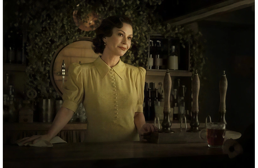 Catherine Zeta-Jones, in "A Visit To Grandpa's," in a vintage yellow dress, stands behind a bar covered with beer taps, surrounded by leafy decor and bottles, conveying a warm, nostalgic atmosphere.