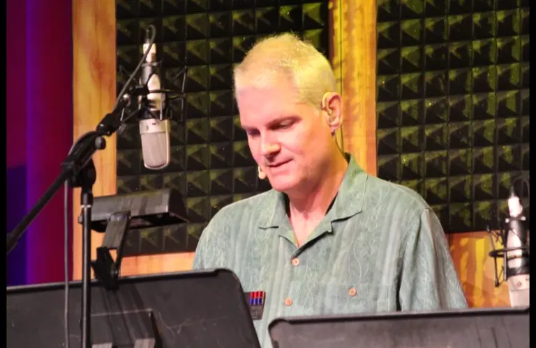 Tom Kane, a person with short blond hair is speaking into a microphone in a recording studio. They wear a green shirt and have a focused expression.