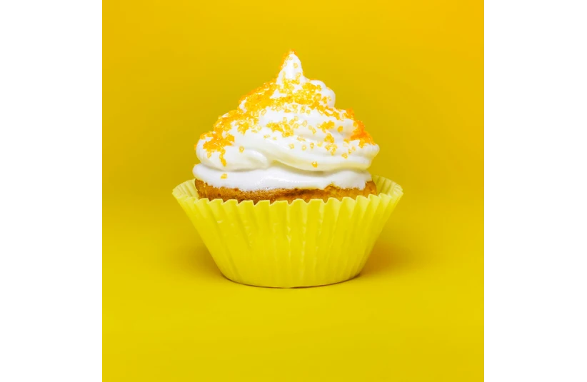 Cupcake with white frosting and orange sprinkles in a yellow wrapper on a bright yellow background, creating a cheerful and vibrant mood.