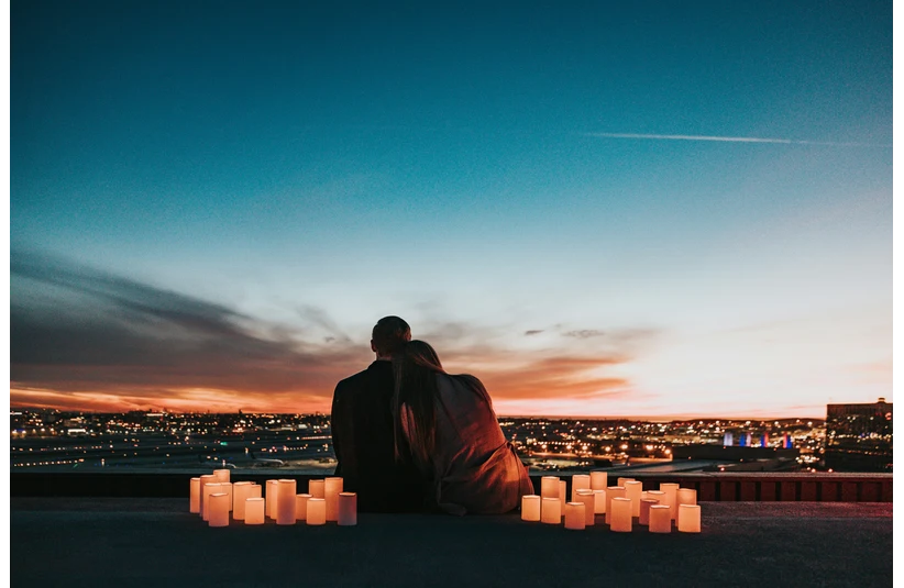 A couple sits on a rooftop at sunset, surrounded by lit candles. They gaze at a vibrant city skyline, creating a romantic and serene atmosphere.