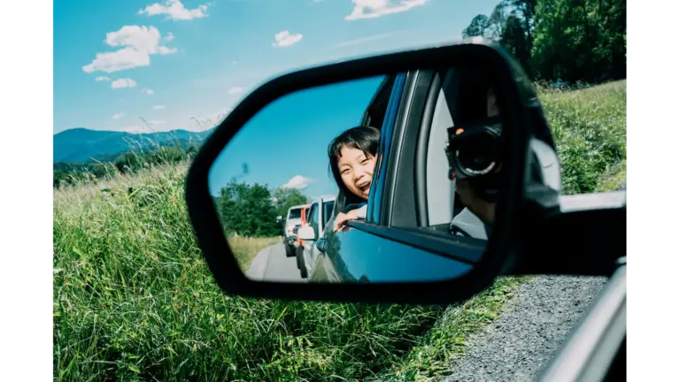 A smiling child looks out of a car’s side‑view mirror during a sunny adventure through lush greenery, capturing the carefree magic of road trips.