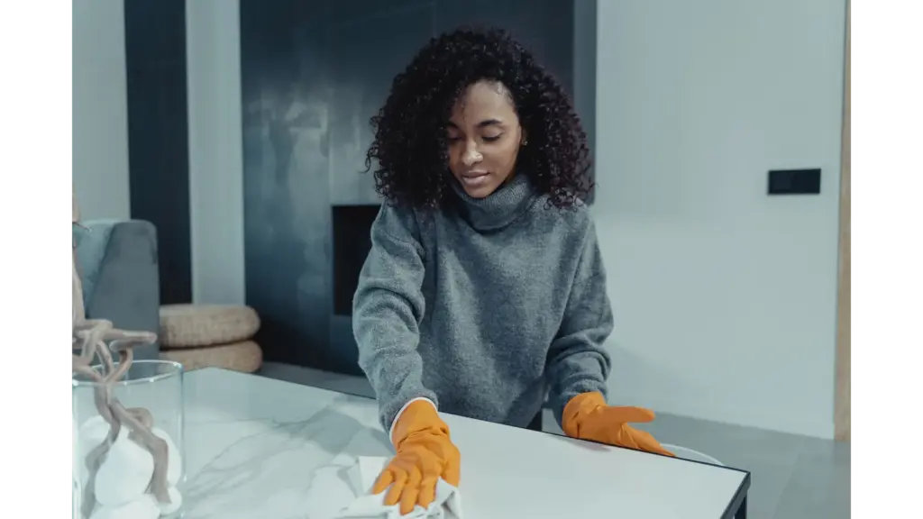 A woman wearing gloves wipes a table in a cozy modern living room, capturing the calm, satisfying rhythm of everyday home cleaning.