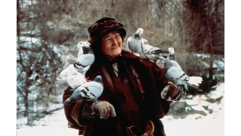 Brenda Fricker in Home Alone 2: Lost in New York (1992) wearing a dark coat and hat, smiling, with several pigeons perched on her arms and shoulders in a snowy outdoor setting.