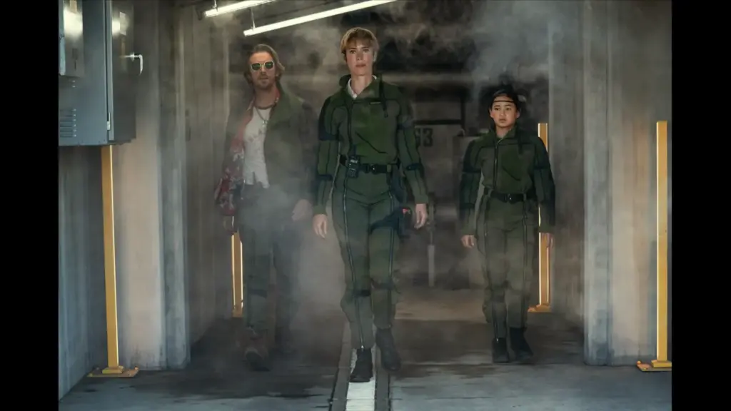 Rebecca Hall, Dan Stevens, and Kaylee Hottle in Godzilla X Kong: The New Empire walk confidently through a smoky corridor. Two wear green tactical gear, while one wears a casual outfit with sunglasses. The mood is intense.