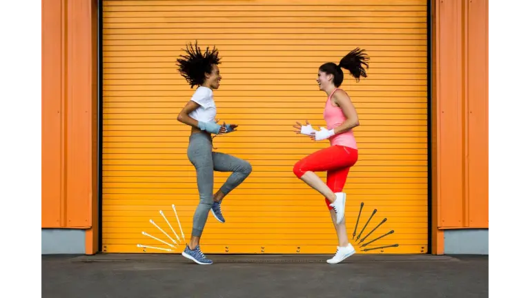 Two healthy women in athletic wear jump playfully in front of a bright orange corrugated metal garage door.