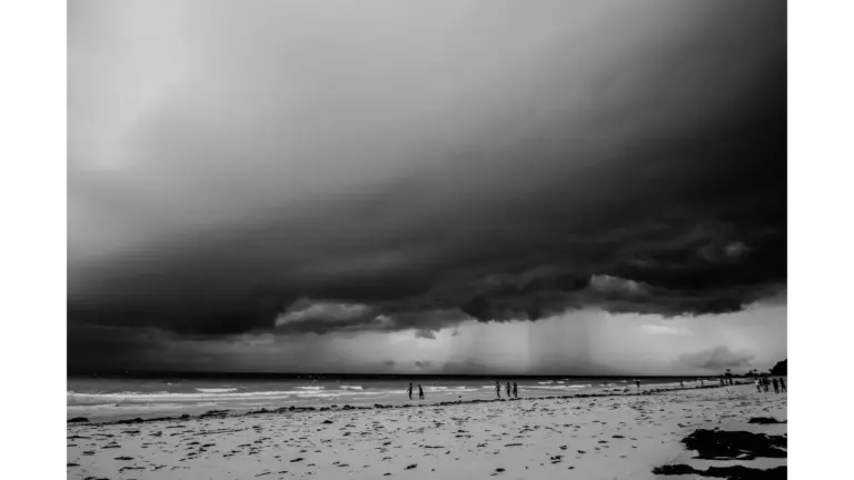 Monochrome view of an impending storm over a beach with people walking along the shore, capturing the dramatic mood of storm travel.