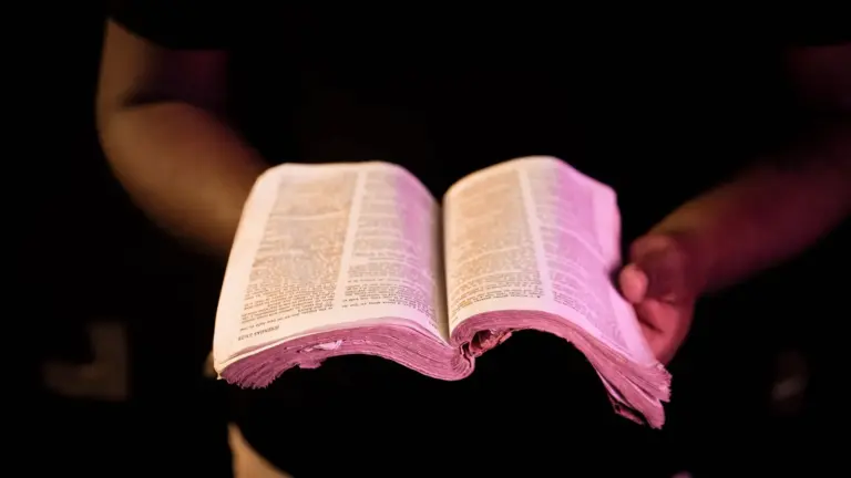 In daily devotional, a man holds an open Bible in a dark-looking background.