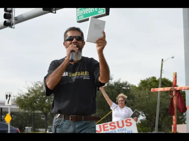 In daily devotional, man boldly preaches in open air setting with Bible held high. 