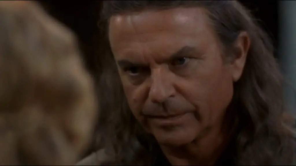 Sam Neill as Merlin in "Merlin's Apprentice" tv series staring intently, conveying determination. His expression is serious, suggesting a tense or dramatic situation. Blurred figure in foreground.