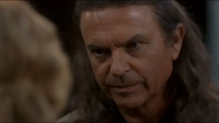 Sam Neill as Merlin in "Merlin's Apprentice" tv series staring intently, conveying determination. His expression is serious, suggesting a tense or dramatic situation. Blurred figure in foreground.
