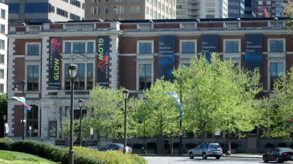 Academy of Natural Sciences of Drexel University -sunlit city street lined with leafy trees and parked cars, facing a brick building with colorful banners; a calm, bustling downtown scene.