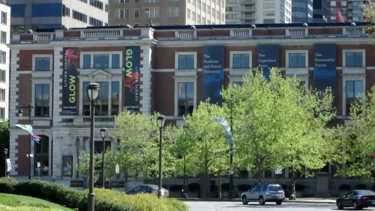 Academy of Natural Sciences of Drexel University -sunlit city street lined with leafy trees and parked cars, facing a brick building with colorful banners; a calm, bustling downtown scene.
