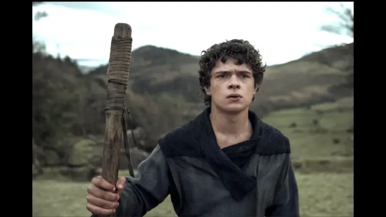 A young man poses in a field, holding a wooden stick, with greenery and trees visible in the distance. Noah Jupe in The Death of Robin Hood - Noah Jupe casted in Avengers: Secret Wars