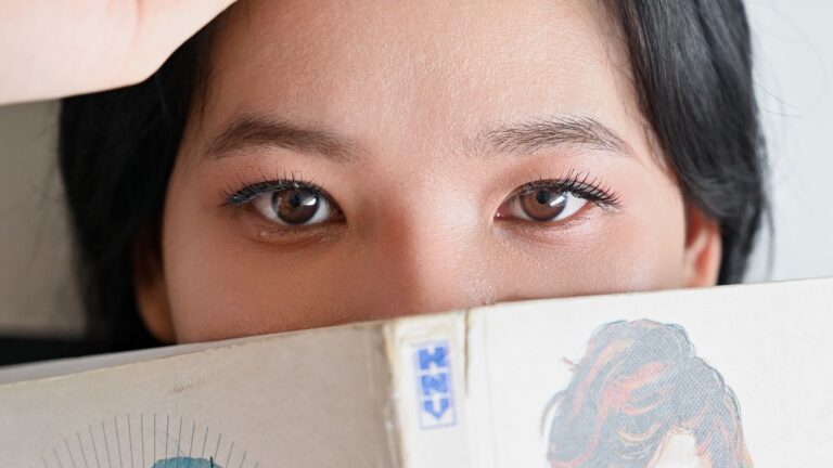 A young woman peeks over the top of an open manga, the kind of shojo romance She Loves Lies Too Much fans are waiting on