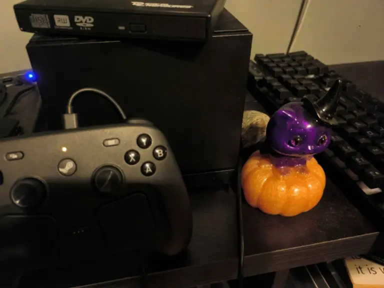 A Steam Machine with Steam Controller sitting on an entertainment stand, a purple cat with black witches hat sitting on an orange pumpkin, a visable keyboard and DVD drive sitting on top.