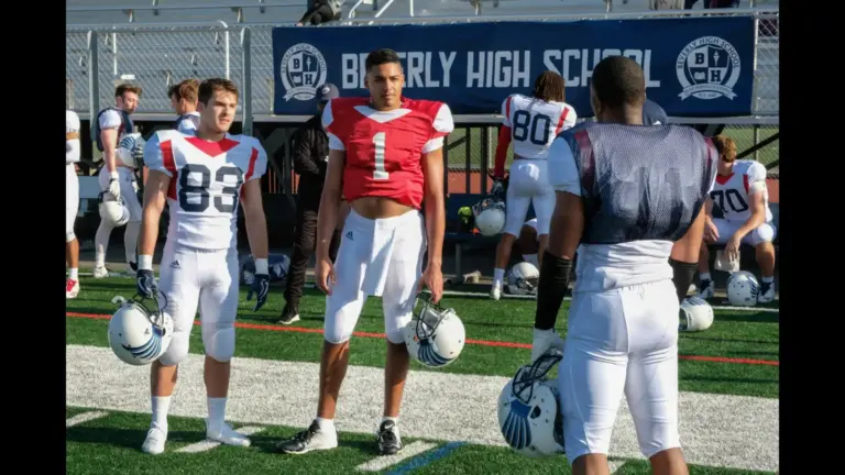 Two football players in white uniforms and helmets stand on a field, looking at a third player wearing a red jersey and a helmet, who is also on the field. A banner in the background reads "BEVERLY HIGH SCHOOL." Cody Christian, Daniel Ezra, and Michael Evans Behling in All American (2018), Asher Adams