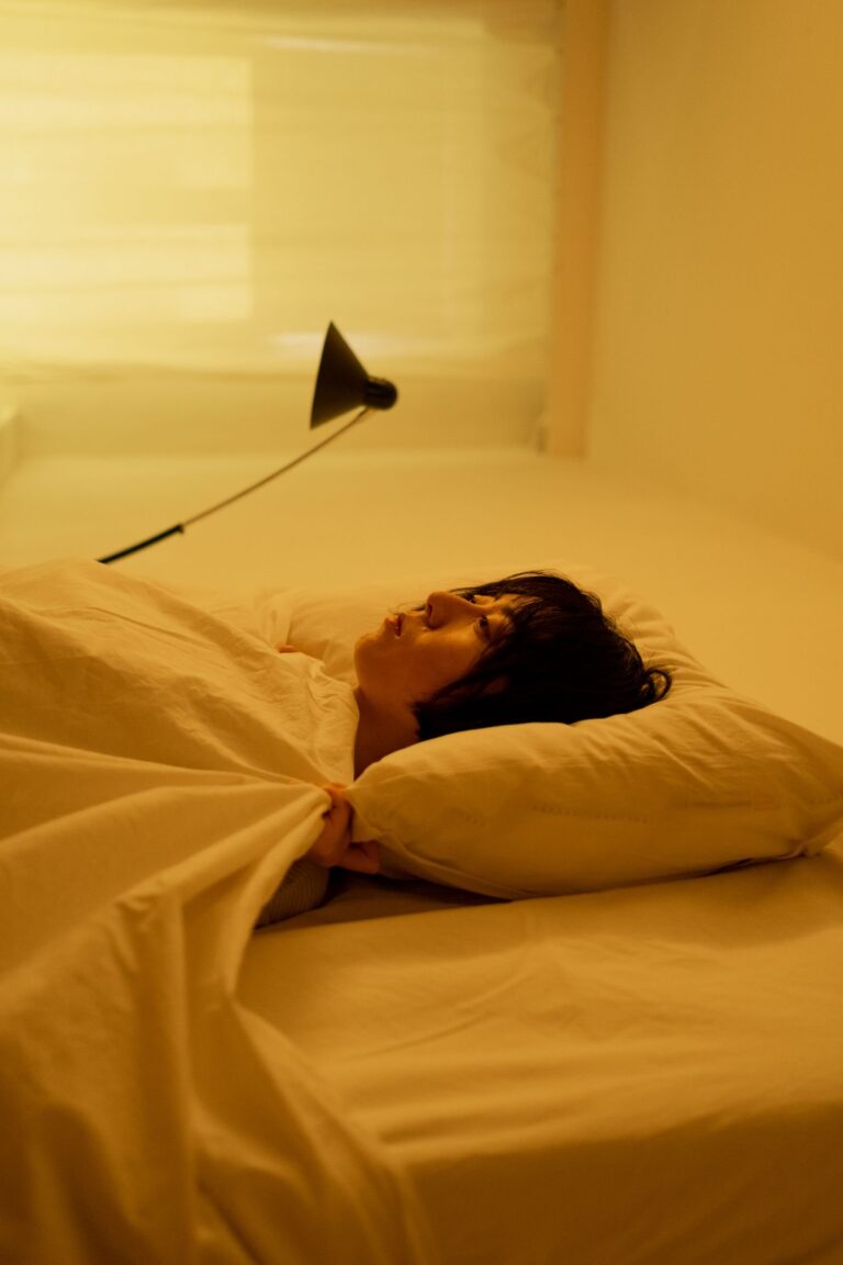 A woman lies in bed under warm lamplight, clutching the covers, as many people try to stop overthinking at night